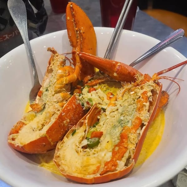 Two halves of baked lobster, topped with cheese and vegetables, served in a white bowl.