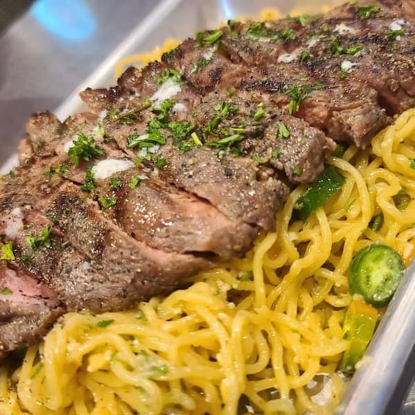 Sliced grilled steak with herbs and seasoning, served over yellow noodles with green vegetables.